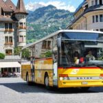 Bus jaune en mouvement sur une rue pavée dans un quartier historique avec des montagnes en arrière-plan