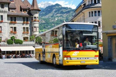 Bus jaune en mouvement sur une rue pavée dans un quartier historique avec des montagnes en arrière-plan