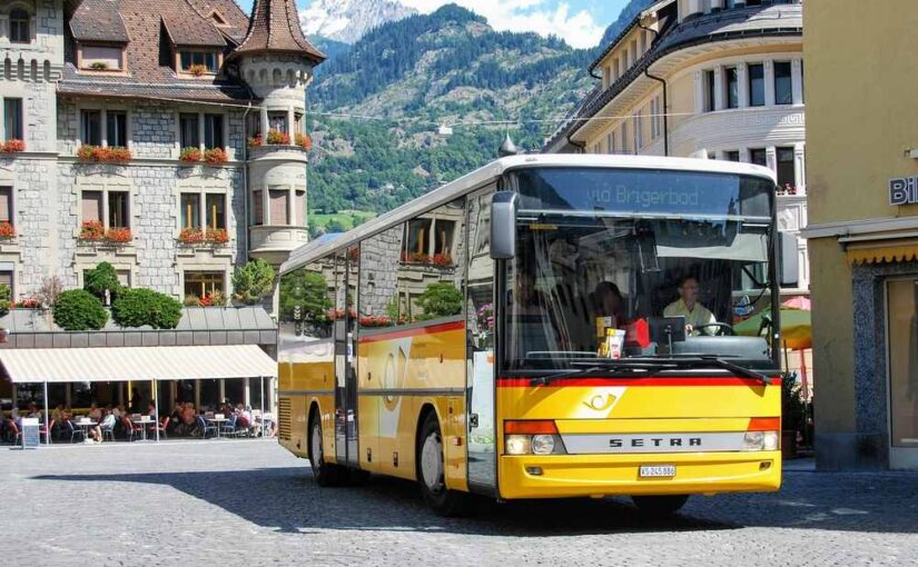 Bus jaune en mouvement sur une rue pavée dans un quartier historique avec des montagnes en arrière-plan